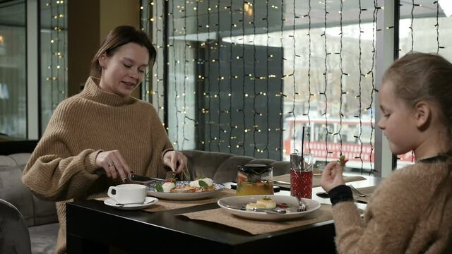 mother and daughter have a delicious meal in a cafe sitting at a table by the window. Family breakfast in the cafe. High quality FullHD footage