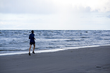 Exercising outside. A jogger in lightweight blue long sleeve t-shirt, shorts and running sneakers trains to marathon, running across the Baltic Sea beach, on cloudy windy rainy early morning