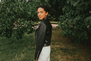 Fashion portrait of attractive confident young natural beauty African American woman with afro hair in black leather jacket posing in nature park in green foliage.
