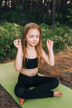 A little preschool girl, a child athlete sits on a green rug in a lotus position, crossing her legs, performing complex sports exercises of gymnastics, trains yoga in the forest. Sports photography.