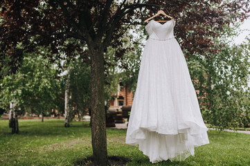 Beautiful white lace dress of the bride hangs on a wooden hanger on a tree in the garden in nature.