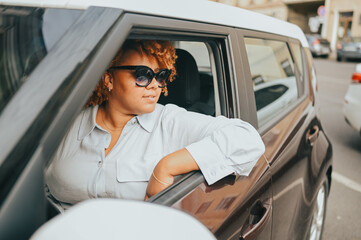 Happy young smiling African American woman red afro haired driver in sun glasses sitting in new brown car, smiling looking at camera enjoying journey. Driving courses and life insurance concept.