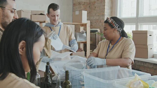 Slowmo tracking shot of team of cheerful multi-ethnic volunteers having conversation while sorting out garbage into recycling containers working together indoors