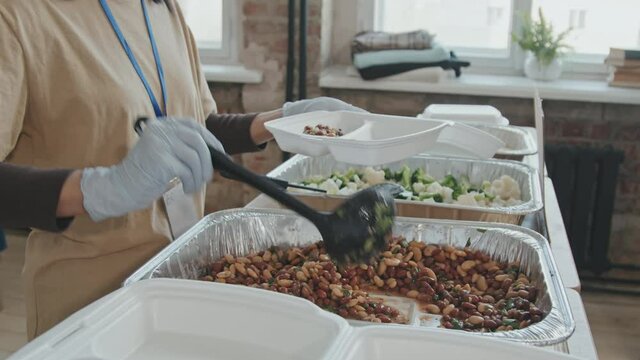Midsection slowmo of unrecognizable volunteer putting food in plastic container for giving homeless and needy people