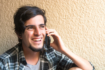 Fototapeta premium Young caucasian black haired man with plaid shirt calling or talking on the phone laughing and smiling holding a smartphone on his hands.