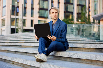 An office worker with laptop on his knees sits on step in front of building of business center.