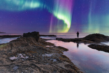 Man’s reflection silhouette as a Green Aurora borealis shimmers over the ocean water - obrazy, fototapety, plakaty