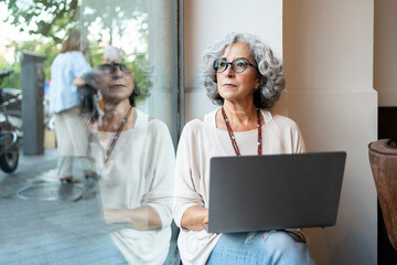Mature woman using laptop and looking through window with thoughtful expression in a coffee shop