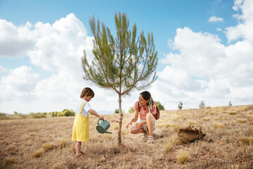 Boy with mother watering lonely tree in field
