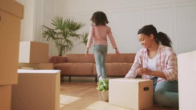 Little girl helping her mom with relocation, bringing her plant and embracing with daddy on sofa, woman unpacking boxes
