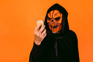 Man dressed in pumpkin latex mask and hooded velvet cape, looks at his mobile phone. Halloween and...