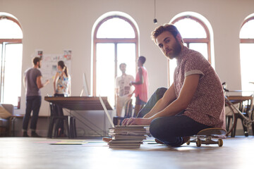 Creative businessman on skateboard working at laptop on floor in open office