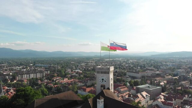 Slow drone flying up with a view of Ljubljana city above the castle with the flag of the city and a national Slovenian flag
