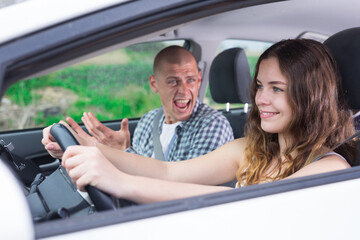 Smiling young girl confidently driving car against background of angry screaming husband sitting in passenger seat