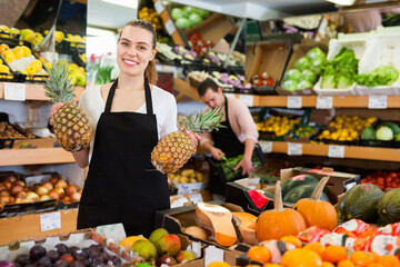 Young happy cheerful positive smiling saleswoman wearing apron showing pineapples on the supermarket