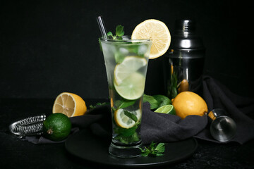 Glass of fresh mojito, shaker, strainer and citrus fruits on dark background