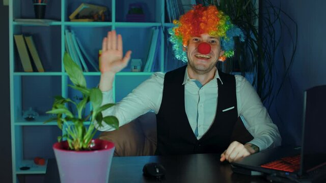 Young man in image of clown waving his hand and looking at camera in office. Funny clown sits in armchair before computer and welcomes. Concept of clown at work.