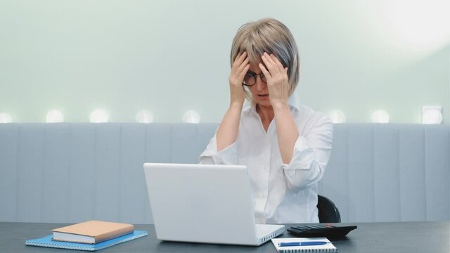 A tense middle-aged elderly business lady, very upset about a big problem or a mistake that she discovered while working at her laptop at home.