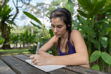 Girl sitting on a wooden bench in the park listening to music on her headphones writes in her diary or notepad. 