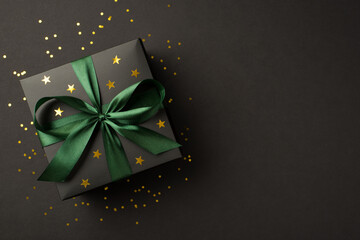 Top view photo of stylish giftbox with green ribbon bow golden stars and sequins on isolated black background with blank space