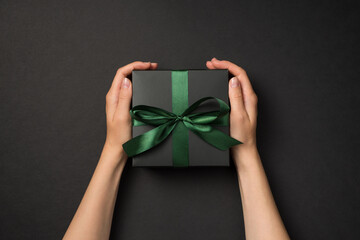 Top overhead first view person photo of girl's hands holding beautiful black present box with silky green ribbon isolated dark backdrop