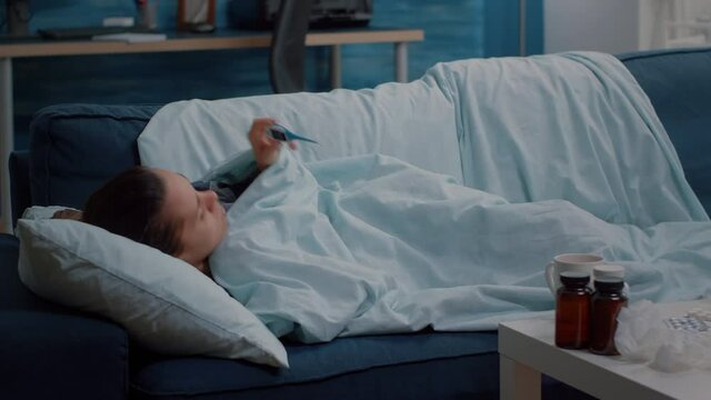 Ill person laying on couch and holding thermometer to check temperature. Sick woman looking at medical intrument, measuring fever for disease diagnosis. Adult with virus symptoms