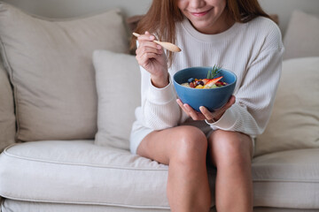 A young asian woman eating healthy blueberry smoothie bowl with mixed fruits topping on sofa at home