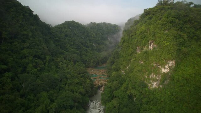Drone aerial hyperlapse of Guatemalan rainforest Semuc Champey National Park and river Rio Cahabon on a cloudy afternoon with light clouds and fog rolling up the green mountain and hillsides. 
