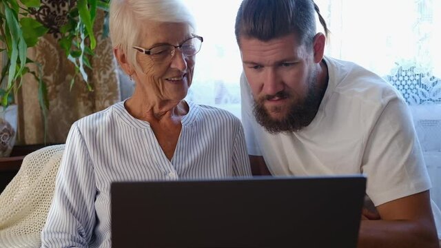 Happy senior mother, adult son sitting in living room at sofa and using laptop trying to make online payment. retirement lifestyle, family spending quality time at home. middle-aged man helping mum