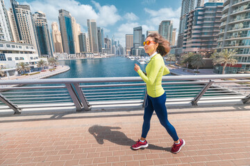 Happy Asian woman in sportswear runs along the Dubai Marina embankment with a smartphone and headphones. Fitness classes and health concept