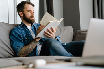 Bearded man writing down notes while working with laptop on sofa