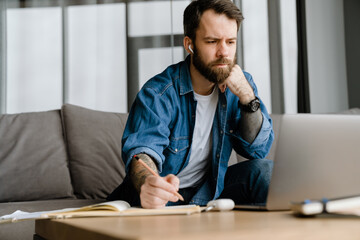 Bearded man writing down notes while making conference call on laptop