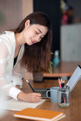 Young beautiful asian woman standing in office and writing note on the table.