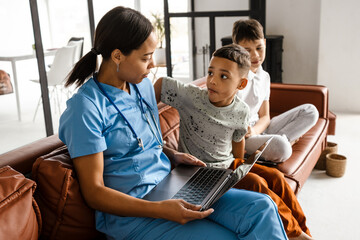 Black woman doctor using laptop while sitting with her sons on couch