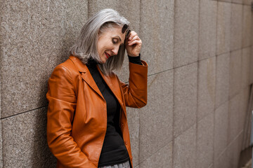 Grey senior woman in leather jacket smiling while leaning on wall