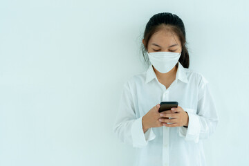 Asian teenage girl wearing white shirt Leaning against a white wall, pressing a cell phone with a medical mask covering his mouth and nose.