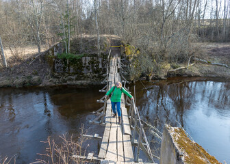 Fototapeta na wymiar spring landscape with a small wild river, bare trees without leaves, old wooden bridge over the river, woman in green jacket on the bridge