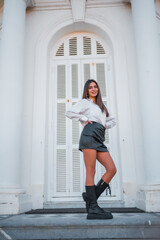 Lifestyle, fashionable pose of a young brunette with a black leather skirt and a shirt on the stairs of a white house