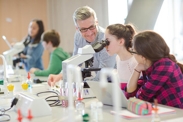 Male teacher helping girl students at microscope in laboratory classroom