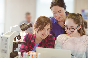 Female teacher and girl students using microscope and laptop, conducting scientific experiment in...