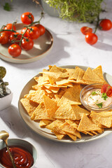 A grey plate with cheese tortilla chips and a chilli cheese dip on a grey surface, cherry tomatoes, ketchup in hard morning light