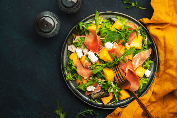 Fresh salad with cantaloupe melon, prosciutto, soft cheese and arugula on blue background, top view, copy space