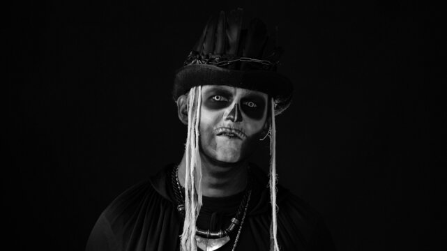 Sinister man, professional skull makeup making faces and ominously smiling with black teeth. Scary guy in thematic carnival costume of Halloween skeleton against black background. Black white image