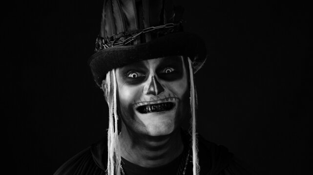 Sinister man with skull makeup opening his mouth and showing dirty black teeth. Portrait of scary guy in thematic carnival costume of Halloween skeleton against black background. Black and white