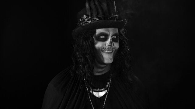 Creepy man with skeleton makeup in top-hat. Guy turning his head and looking at camera, trying to scare. Voodoo rituals. Baron Saturday. Halloween thematic party. Black background. Black and white