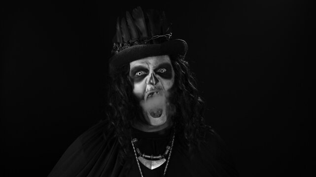 Sinister man with horrible Halloween skeleton makeup in costume with top-hat exhaling cigarette smoke from his mouth and smiling on black background. Horror theme. Day of The Dead. Black and white