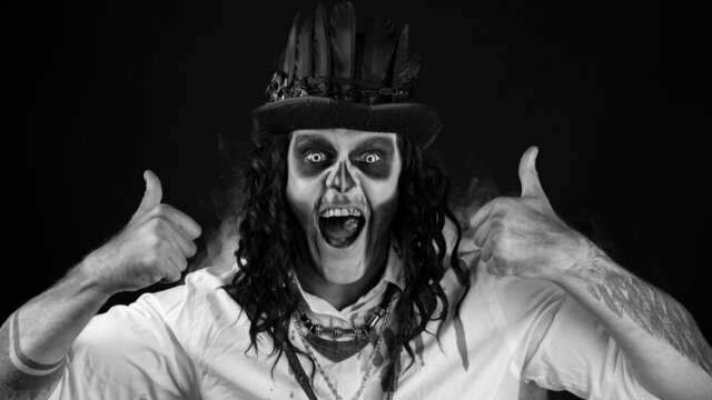 Frightening smiling man in skeleton Halloween cosplay costume. Guy in creepy skull makeup making faces, looking at camera, showing thumbs up gesture. Day of The Dead. Black background. Black and white