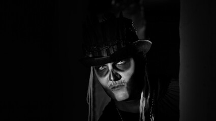 Fototapeta na wymiar Creepy man with horrible Halloween skeleton makeup in costume with top-hat appears from dark corner, trying to scare. Horror theme. Shooting in dark parking hallway. Black and white image
