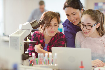 Female teacher and girl students using microscope and laptop, conducting scientific experiment in laboratory classroom