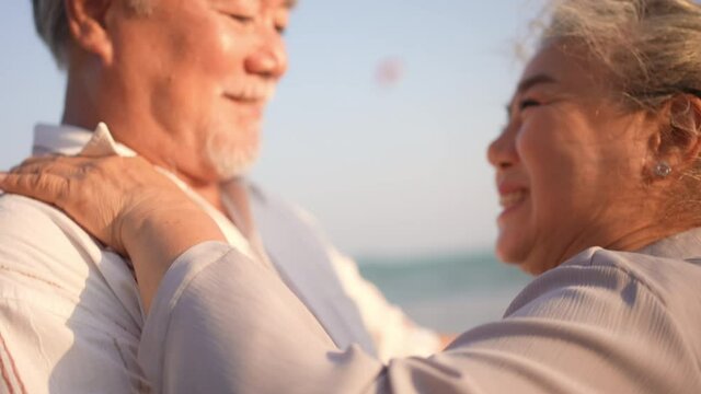 Happy Asian family senior couple holding hand and dancing on the beach together at summer sunset. Elderly retired husband and wife relax and enjoy romantic outdoor holiday vacation with happiness